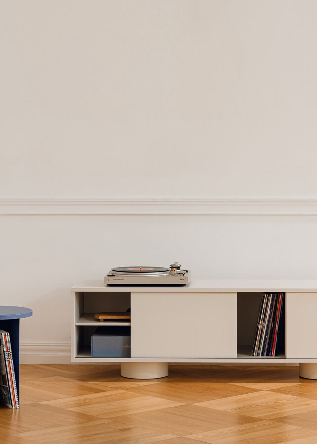 Svetlosivý TV stolík s odsunutými dvierkami v miestnosti na drevenej podlahe pred bielou stenou.