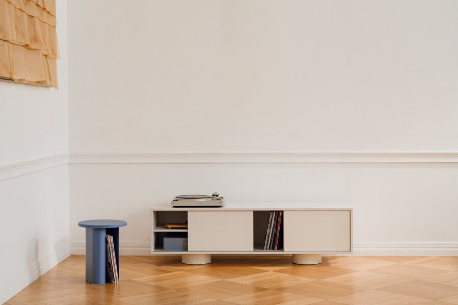 Svetlosivý TV stolík v miestnosti na drevenej podlahe pred bielou stenou, vedľa modrého príručného stolíka.