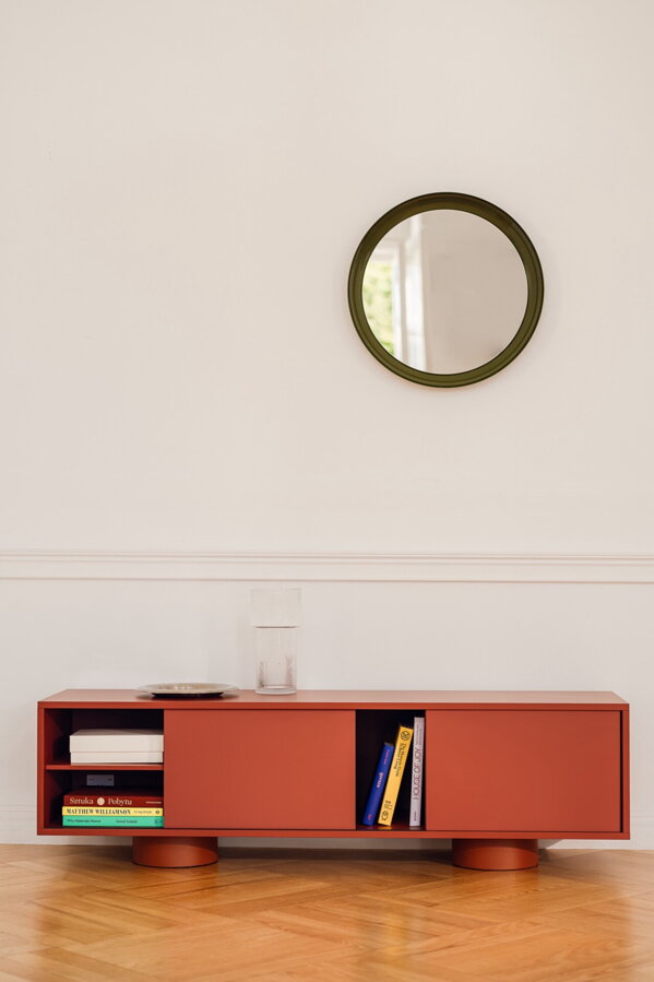 Červenohnedý stolík pod televízor Taro čiernymi oceľovými nohami pri bielej stene pod okrúhlym malým nástenným zrkadlom.
