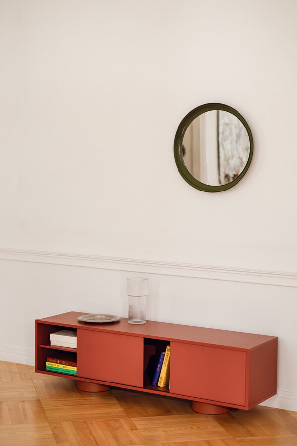 Červenohnedý stolík pod televízor Taro čiernymi oceľovými nohami pri bielej stene pod okrúhlym malým nástenným zrkadlom.