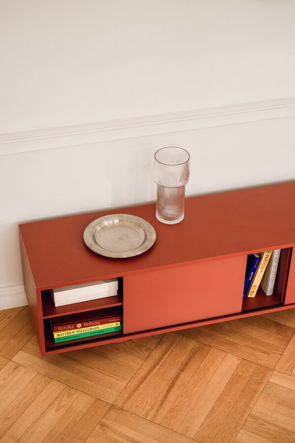 Červenohnedý stolík pod televízor s odsunutými dvierkami, kde v policiach sú poukladané knihy.