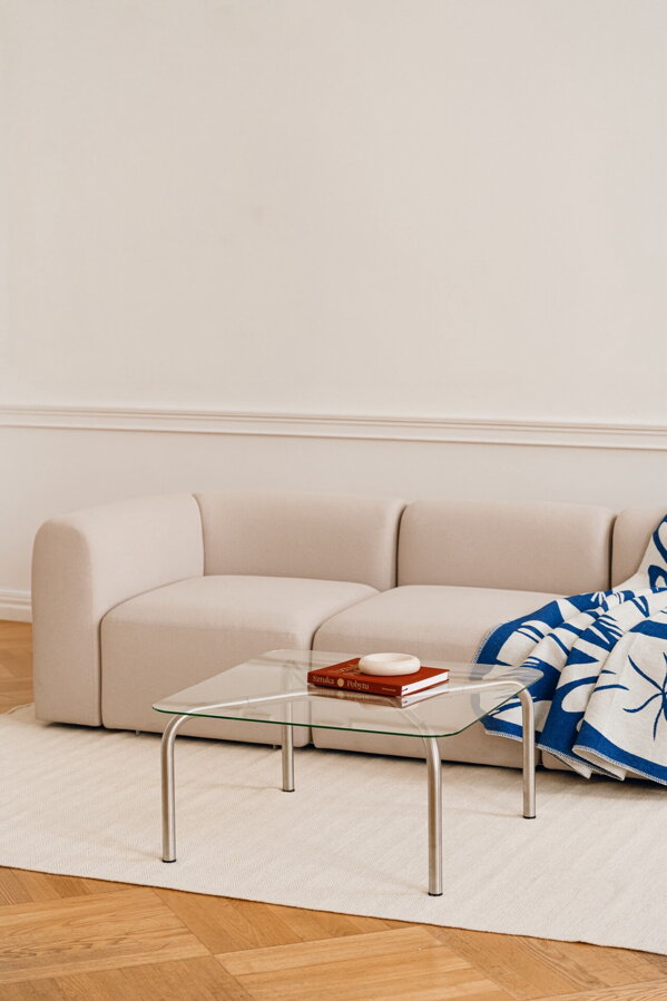 Štvorcový konferenčný stolík Uku so sklenenou doskou a oceľovými nohami na béžovom koberci pred béžovou pohovkou.