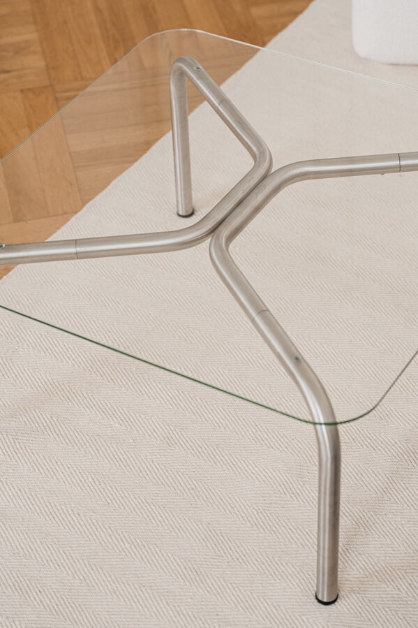 Štvorcový konferenčný stolík Uku so sklenenou doskou a oceľovými nohami na béžovom koberci.