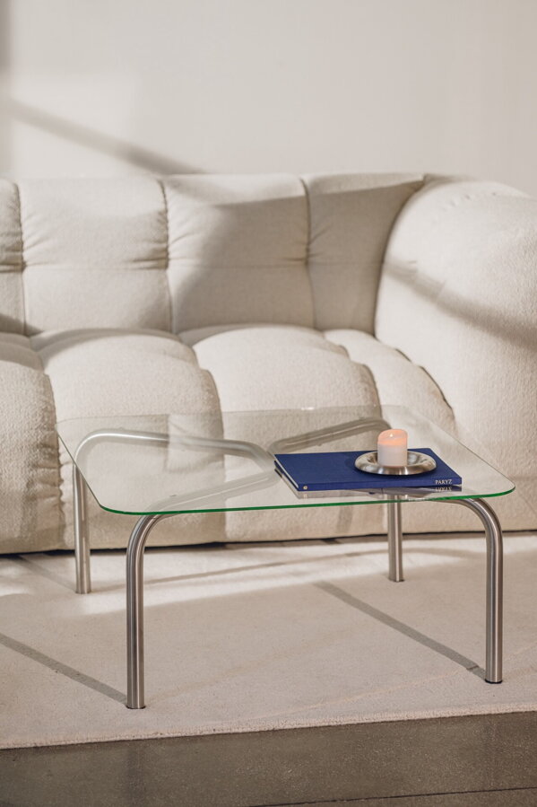 Štvorcový konferenčný stolík Uku so sklenenou doskou a oceľovými nohami na béžovom koberci pred béžovou pohovkou.