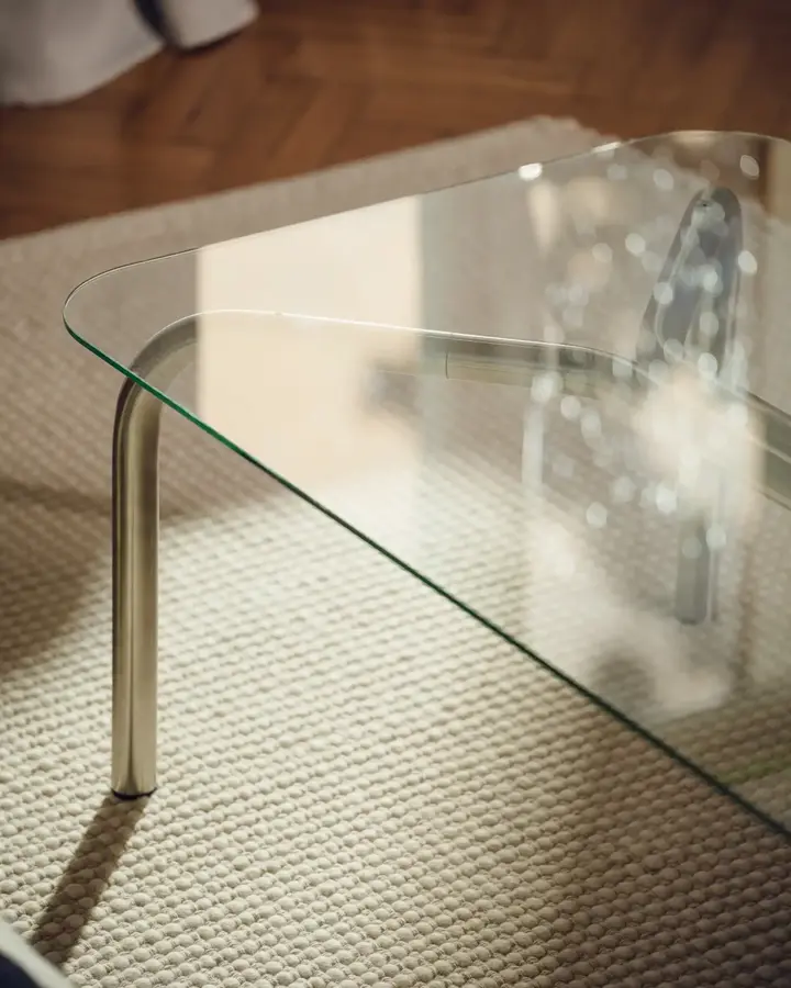 Sklenený obdĺžnikový konferenčný stolík s oceľovými nohami, na bielom koberci.