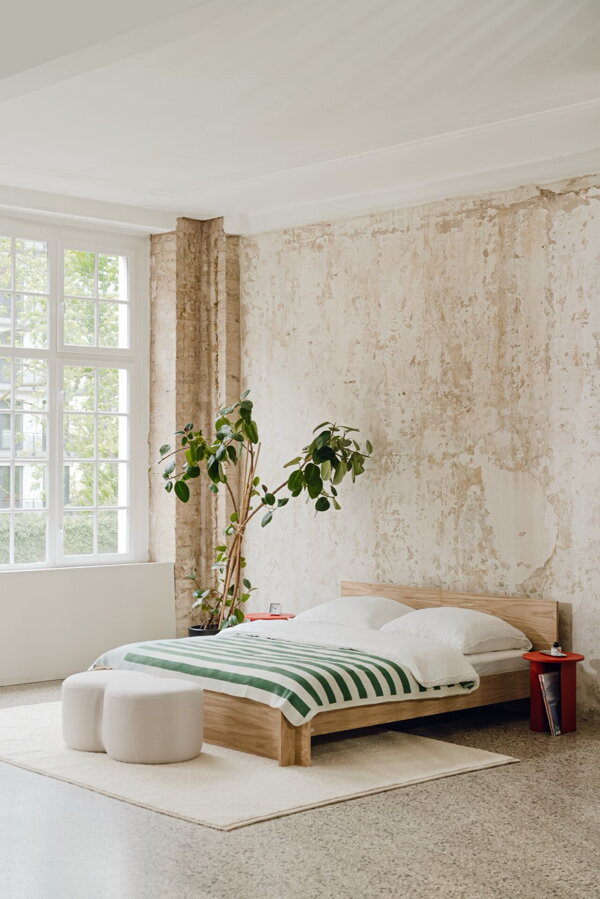 Masívna dubová manželská posteľ prírodnej farby vo dne vo svetlej spálni s veľkým bielym oknom.