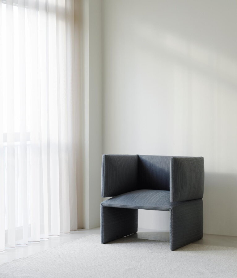 Čalúnené sivé kreslo Fold Lounge Chair pred veľkým oknom na bielom koberci v bielej miestnosti.