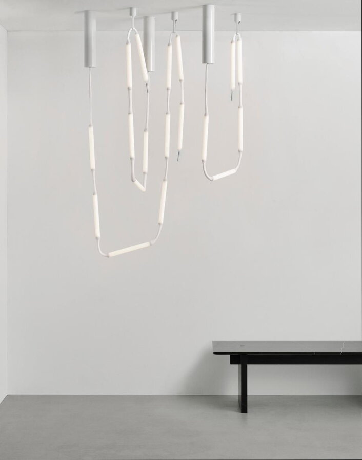 Lampa Reb ako svetelná reťaz visiaca zo stropu v nepravidelných oblúkoch nad čiernym stolíkom vedľa bielej steny.