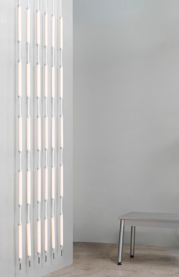 Lampa Reb ako svetelný záves na bielej stene vedľa sivého príručného stolíka/stoličky.