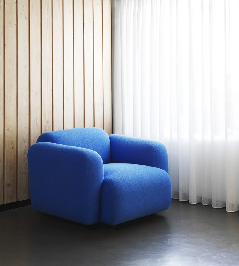 Modré čalúnené kreslo Swell Armchair vedľa béžovej steny pred oknom s bielou záclonou.