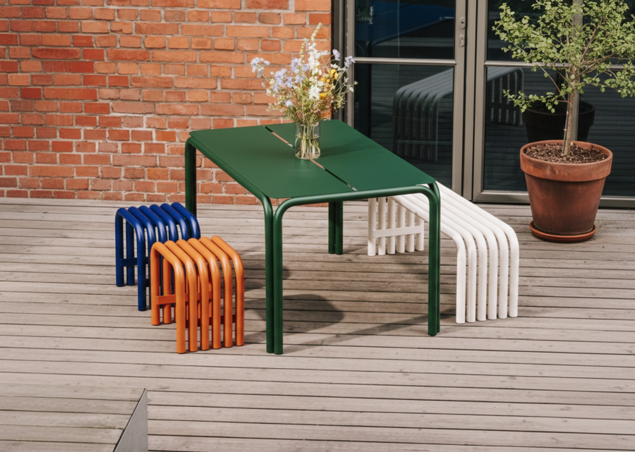 Zelený stôl, oranžová a modrá stolička a biela lavica z galvanizovanej ocele na drevenej terase domu.